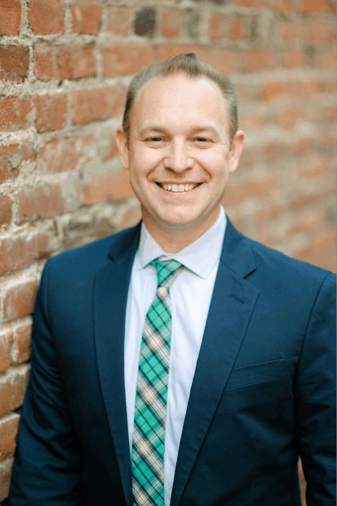 A white man in a navy suit and green tie standing inf front of a brick wall.
